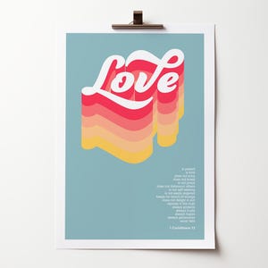 May include: A rectangular wall art print on a muted blue background featuring the word Love in a layered, 3D-style typography with white, red, orange, and yellow gradients. Below the text is small white printed scripture from 1 Corinthians 13.