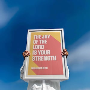 K&ouml;nnte beinhalten: Ein gerahmtes Poster mit dem Text "THE JOY OF THE LORD IS YOUR STRENGTH" in Wei&szlig;, vor einem gelben und korallenfarbenen Hintergrund. Der Text "Nehemiah 8:10" befindet sich unten. Das Poster wird vor einem hellblauen Himmel gehalten.