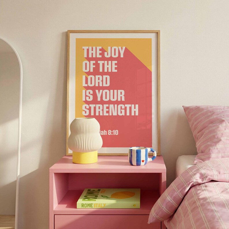 K&ouml;nnte beinhalten: Ein gerahmter Druck mit dem Text "THE JOY OF THE LORD IS YOUR STRENGTH" in Wei&szlig; auf rotem und gelbem Hintergrund. Eine wei&szlig;e und gelbe Lampe, eine blau-wei&szlig;e Tasse und ein Buch stehen auf einem rosa Nachttisch neben einem Bett mit rosa gestreifter Bettw&auml;sche.