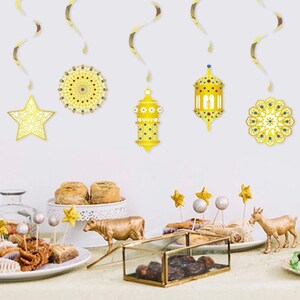 May include: Gold Ramadan party decorations featuring a star, lanterns, and geometric designs. The decorations are hanging from the ceiling and there are gold and white plates with food on a table below.