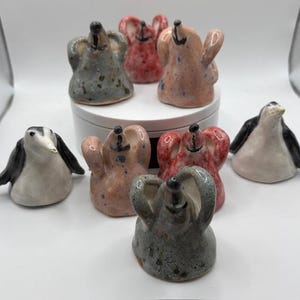 Peut inclure: Ensemble de figurines en céramique, dont des pingouins et des créatures ressemblant à des éléphants. Les figurines sont de différentes couleurs, notamment grises, roses et rouges, avec des finitions mouchetées et vernissées. Les pingouins sont noirs et blancs.