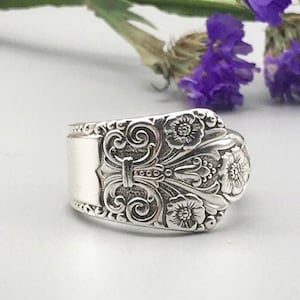 May include: A silver ring with an ornate floral and scroll design. The ring features a rectangular face with a central vertical bar and is surrounded by detailed embellishments. The ring is set against a soft, neutral background with purple flowers.