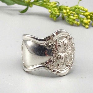 May include: A silver ring crafted from a spoon, featuring a floral design on the face. The ring has a wide band and a polished finish. The ring is a unique piece of jewelry.