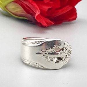 May include: A silver ring crafted from a spoon, featuring ornate floral and leaf detailing. The ring's band is wide, and the spoon's bowl forms the face of the ring. A red flower is in the background.