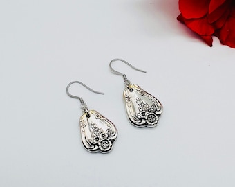 May include: A pair of silver-colored drop earrings. Each earring features a floral design and a hook closure. The earrings are teardrop-shaped and have a vintage aesthetic. The background is white with a red flower in the upper right corner.