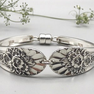 May include: A silver bracelet crafted from repurposed silverware. The bracelet features floral designs on the spoon bowls and a magnetic clasp. The bracelet is a unique piece of jewelry, perfect for adding a touch of vintage charm to any outfit.