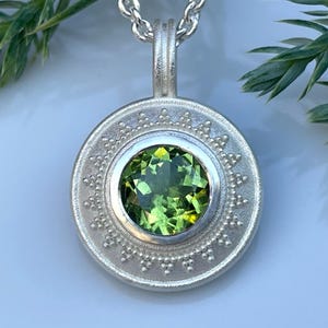 Può includere: Una collana con pendente in argento con un pendente rotondo e ornato con una gemma verde brillante al centro. Il pendente è appeso a una delicata catena. Lo sfondo include fogliame verde.