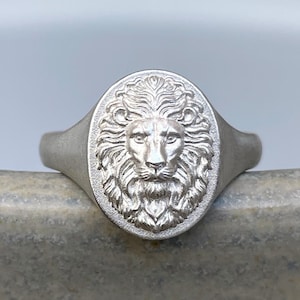May include: A silver-toned signet ring featuring an oval face with a detailed, embossed lion head design. The metal has a matte, brushed finish, and the lion's mane and facial features are clearly defined in the center of the ring's face.