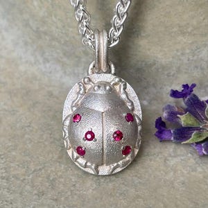 May include: Silver ladybug pendant necklace with red gemstone accents. The oval pendant features a detailed ladybug design with six red gemstones. The necklace is displayed on a neutral background with a sprig of lavender.