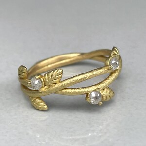 May include: A gold ring with a leaf and vine design. The ring features three small, clear gemstones. The band has a textured, matte finish. The ring is set against a light grey background.