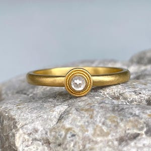 Op de afbeelding: Een gouden ring met een enkele, kleine, ronde, heldere edelsteen in een ronde zetting met een korrelige rand. De band heeft een gladde, matte afwerking en de centrale zetting is verhoogd, met een heldere, gefacetteerde steen in het midden.