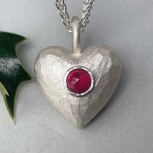 Op de afbeelding: Zilveren hartvormige hanger met een getextureerd oppervlak en een rode edelsteen in het midden, hangend aan een zilveren ketting. Een groen hulstblad staat op de achtergrond. Een romantisch accessoire.