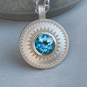 Può includere: Un ciondolo rotondo in argento con una gemma blu brillante sfaccettata al centro. Il ciondolo presenta una superficie opaca e testurizzata con un bordo decorativo a perline che circonda la pietra ed è appeso a una catena a maglie d'argento.