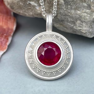 Könnte beinhalten: Eine silberne Anhängerkette mit einem runden, facettierten, tiefroten Edelstein. Der Anhänger hat einen detaillierten, dekorativen Rand. Die Halskette ist ebenfalls aus Silber. Der Hintergrund umfasst eine Muschel und einen grauen Stein.