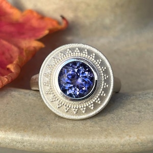 May include: A silver ring with a large, round, deep blue gemstone at the center. The ring's setting is circular with a detailed, textured pattern around the gem. A red and orange leaf rests nearby.