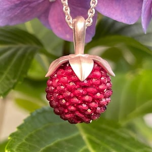 May include: A silver pendant shaped like a raspberry, featuring a textured, deep red, faceted surface. The pendant is attached to a silver chain by a smooth silver stem and leaf cap. The design mimics the appearance of a fresh berry fruit.