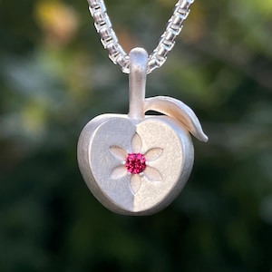 Puede incluir: Un colgante de manzana en plata mate suspendido de una cadena de caja a juego. La manzana presenta un detalle central tallado en forma de estrella con una pequeña piedra preciosa redonda de color rosa vibrante. El metal tiene un acabado suave.