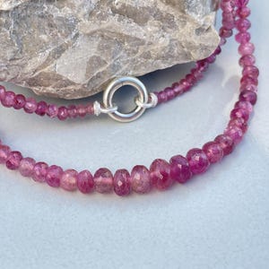 Könnte beinhalten: Eine mehrreihige Halskette mit kleinen, facettierten, rosa Edelsteinen. Die Halskette wird mit einem silbernen Verschluss gesichert. Die Edelsteine variieren in Größe und Form und erzeugen ein strukturiertes Aussehen. Der Hintergrund umfasst einen grauen Felsen und eine hellblaue Oberfläche.