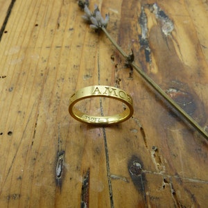 K&ouml;nnte beinhalten: Ein goldener Bandring mit der Gravur "AMOR" um das Band. Der Ring liegt auf einer Holzoberfl&auml;che.