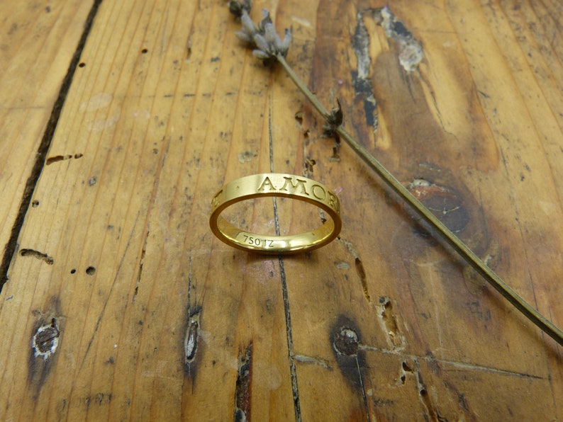 K&ouml;nnte beinhalten: Ein goldener Bandring mit der Gravur "AMOR" um das Band. Der Ring liegt auf einer Holzoberfl&auml;che.
