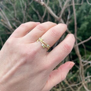 May include: A gold ring with a vine-like design, adorned with small, clear gemstones. The ring is on a finger, set against a blurred backdrop of green foliage and brown branches.