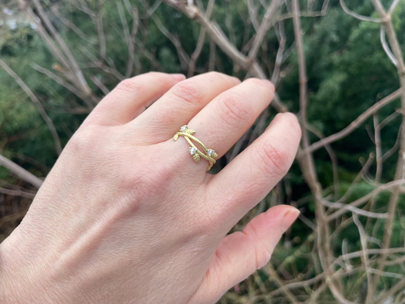 May include: A gold ring with a vine-like design, adorned with small, clear gemstones. The ring is on a finger, set against a blurred backdrop of green foliage and brown branches.