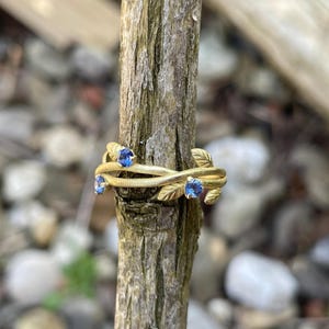 May include: A gold ring with a delicate design of intertwined branches and blue gemstones. The ring is displayed on a wooden branch.
