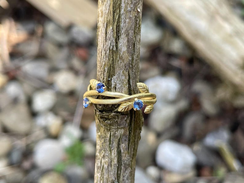 May include: A gold ring with a delicate design of intertwined branches and blue gemstones. The ring is displayed on a wooden branch.