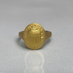May include: A gold ring featuring a raised scarab beetle design on the top. The ring is oval shaped and has a smooth band.