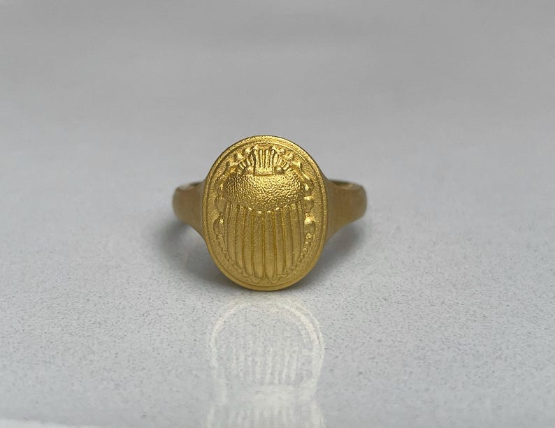 May include: A gold ring featuring a raised scarab beetle design on the top. The ring is oval shaped and has a smooth band.