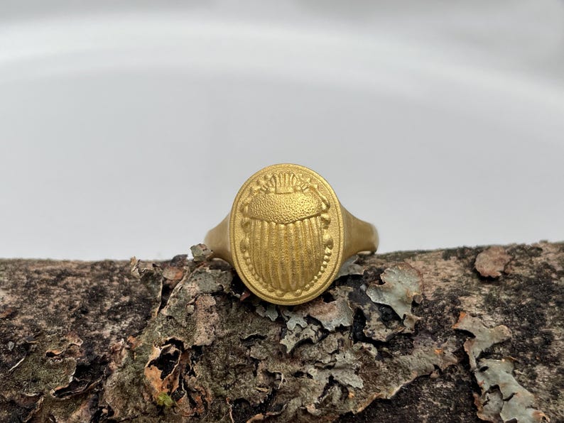 May include: A gold ring with an oval-shaped design featuring a raised, detailed image of a scarab beetle.