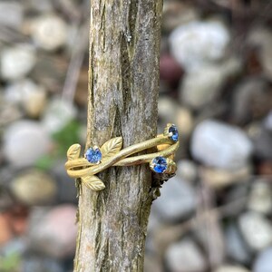 May include: A gold ring with a delicate vine design and three blue sapphires. The ring is on a wooden branch.