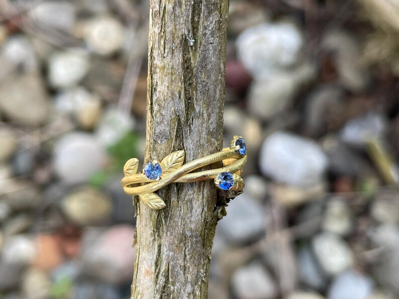 May include: A gold ring with a delicate vine design and three blue sapphires. The ring is on a wooden branch.