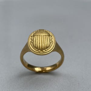 May include: A gold ring featuring a raised scarab beetle design. The ring is a simple band with a round bezel.