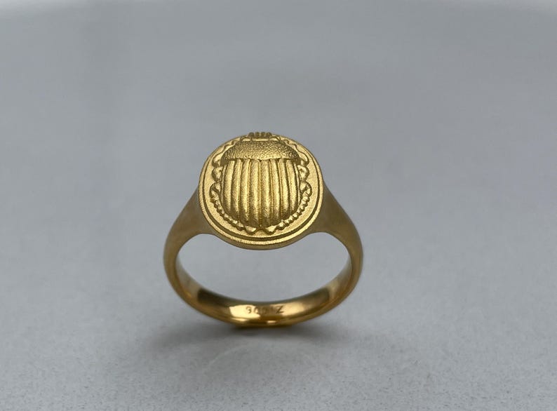 May include: A gold ring featuring a raised scarab beetle design. The ring is a simple band with a round bezel.