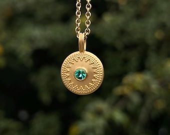 May include: A gold-toned circular pendant necklace featuring a central round teal gemstone. The pendant surface displays a textured border with a repeating beaded pattern and a zigzag design. It hangs from a matching gold-toned metal chain.