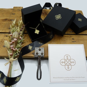 May include: A gold necklace with a geometric pendant in a black gift box. The box is surrounded by black ribbon with a gold logo. A card with a quote by Leonardo da Vinci is also included. "Wiewiel Sch&ouml;nheit empf&auml;ngt das Herz durch die Augen. - Leonardo da Vinci -"