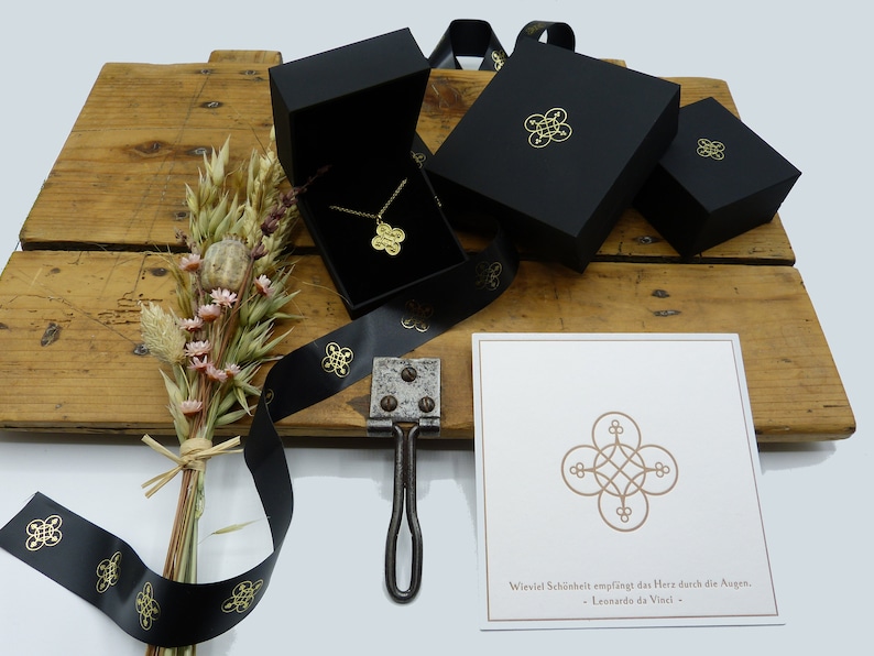 May include: A gold necklace with a geometric pendant in a black gift box. The box is surrounded by black ribbon with a gold logo. A card with a quote by Leonardo da Vinci is also included. "Wiewiel Sch&ouml;nheit empf&auml;ngt das Herz durch die Augen. - Leonardo da Vinci -"