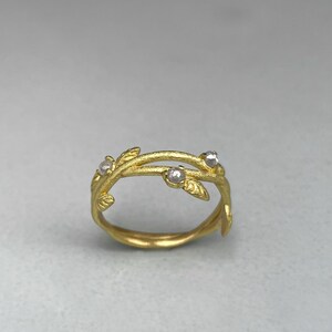 May include: A gold ring with a vine-like design. The ring has small, clear gemstones and leaf details. The band is smooth and the design is inspired by nature. The ring is a piece of jewellery.