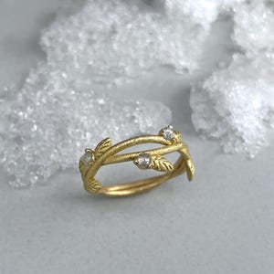 May include: A gold ring with a botanical design. The ring has a vine-like structure with tiny leaves and three clear gemstones. The ring is set against a soft, neutral background, emphasising its intricate details.