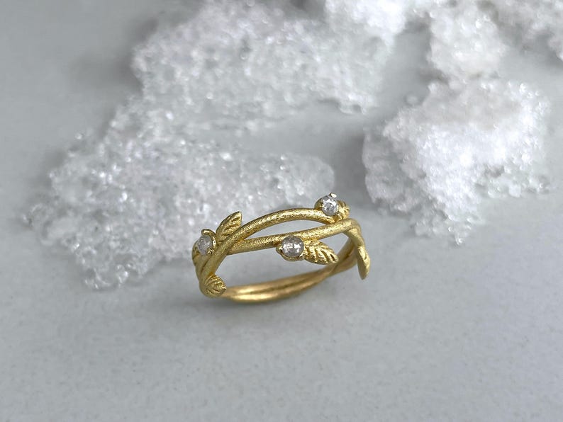 May include: A gold ring with a botanical design. The ring has a vine-like structure with tiny leaves and three clear gemstones. The ring is set against a soft, neutral background, emphasising its intricate details.
