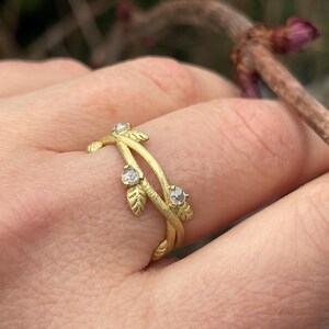 May include: A gold ring with a leaf and vine design, set with three clear gemstones. The ring is worn on a finger. The background is blurred, showing a branch and leaves.