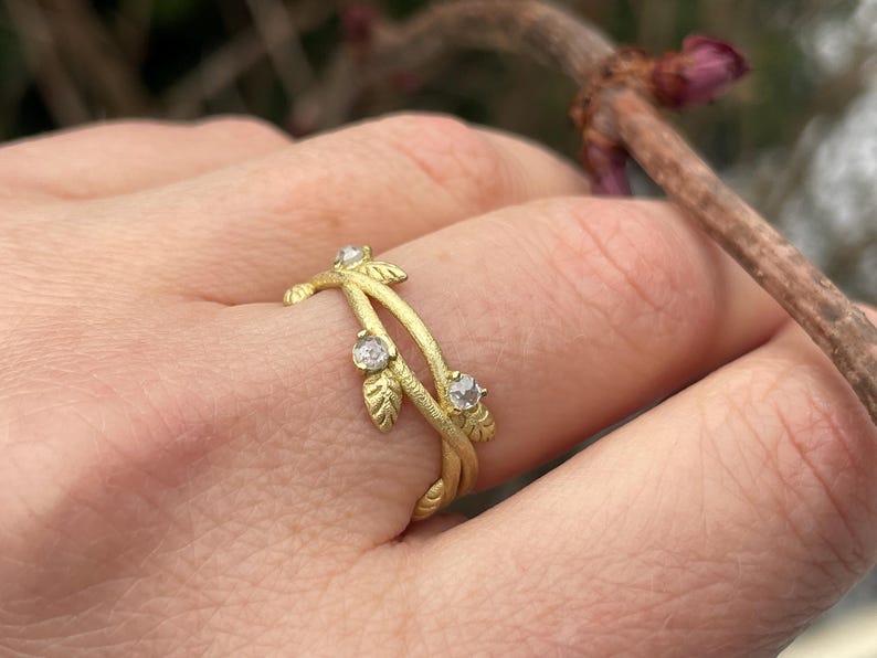 May include: A gold ring with a leaf and vine design, set with three clear gemstones. The ring is worn on a finger. The background is blurred, showing a branch and leaves.