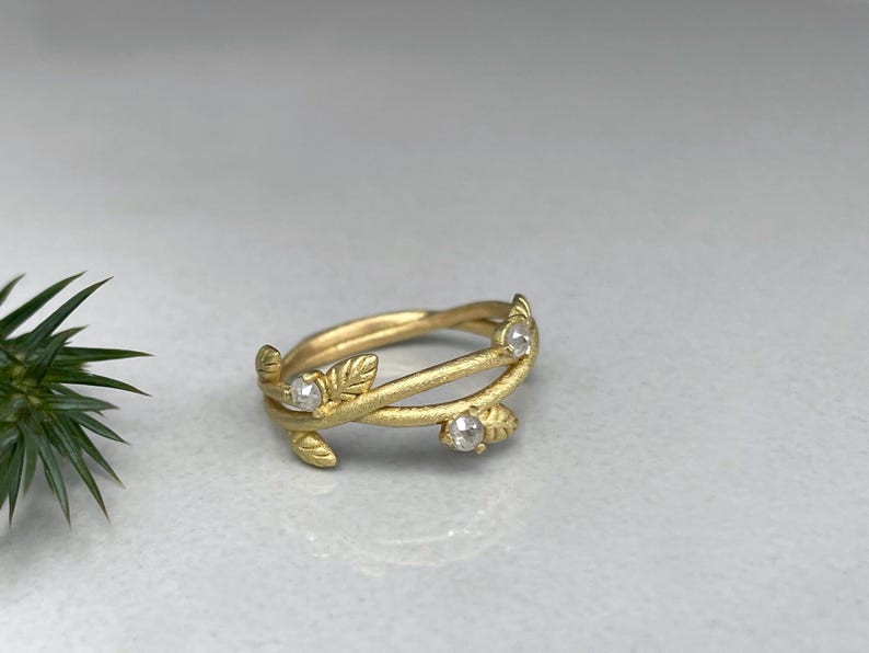 May include: A gold ring with a leaf and vine design. The ring features three small, clear gemstones. The band has a textured, matte finish. The ring is set against a light grey background.