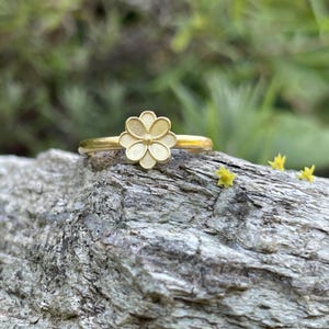 May include: A gold ring with a white flower design. The ring is sitting on a piece of wood.