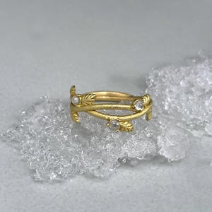 May include: A gold ring with a botanical design. The ring has intertwined bands with small leaf motifs and three clear gemstones. The ring is presented on a textured, light-coloured surface.