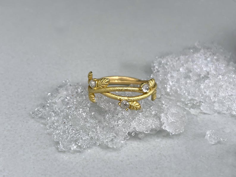 May include: A gold ring with a botanical design. The ring has intertwined bands with small leaf motifs and three clear gemstones. The ring is presented on a textured, light-coloured surface.