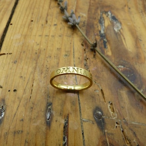 K&ouml;nnte beinhalten: Ein goldener Bandring mit der Inschrift "OMNIA" um die Band.