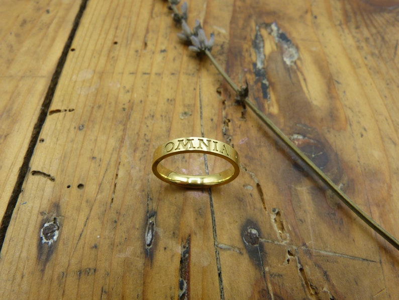 K&ouml;nnte beinhalten: Ein goldener Bandring mit der Inschrift "OMNIA" um die Band.