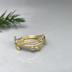 May include: A gold ring with a leaf and vine design. The ring has three small, clear gemstones. The leaves and vines wrap around the band. The ring is on a light grey surface.